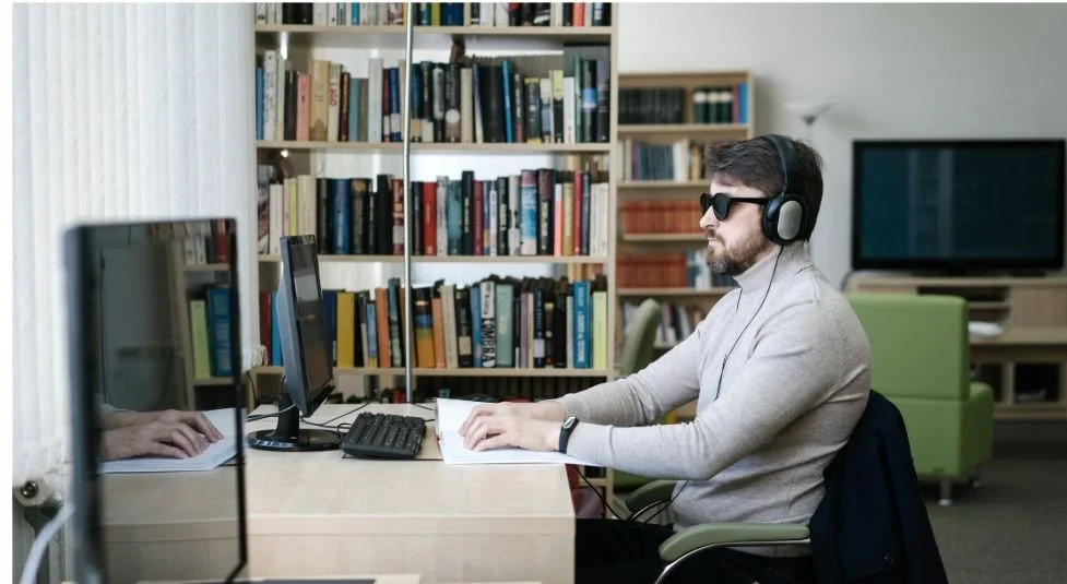 Una persona utiliza una computadora y auriculares en un espacio de trabajo de una biblioteca.